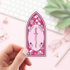 Pink stained glass-style sticker with a sword and roses held by a hand on a light background.