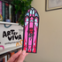 Stained glass-style bookmark with a rose design held by a hand in front of a bookshelf with &