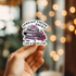 Hand holding a sticker with text and graphics against a blurred background with lights