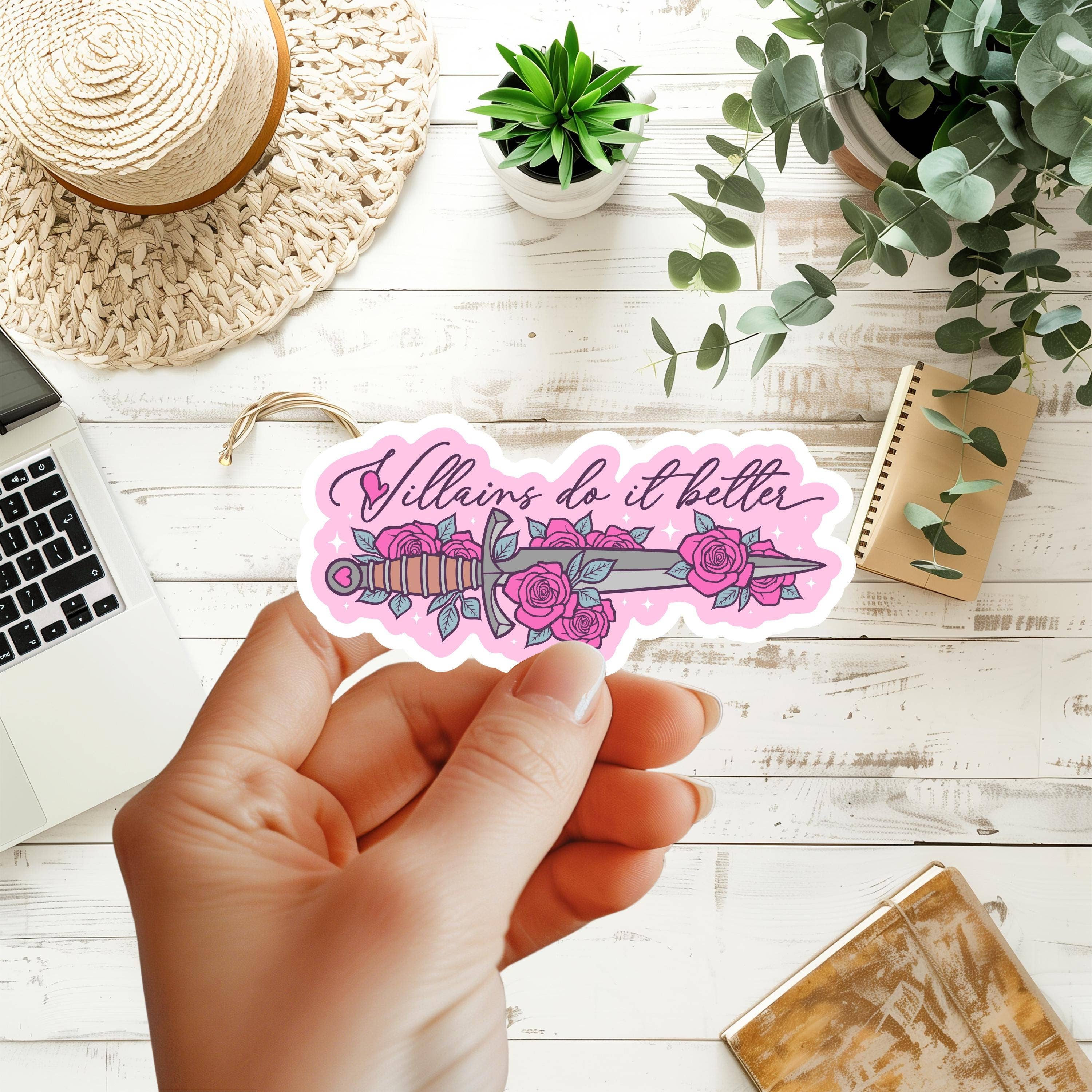 A hand holds a die-cut sticker featuring a pink and purple dagger surrounded by roses with the phrase &