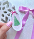 A hand with pink nail polish holds a small oval ornament or sticker featuring a green Christmas tree, white scalloped border, and pink bow, against a glittery light background with a purple ribbon and a white snowflake decoration.