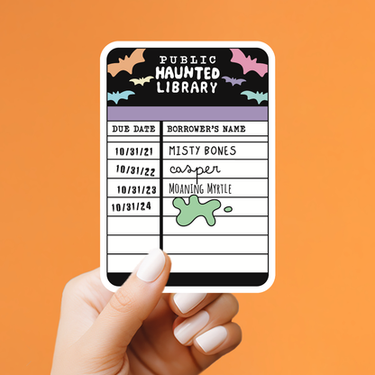 A hand with white manicured nails holds a rectangular sticker against a solid orange background. The sticker is designed like a &
