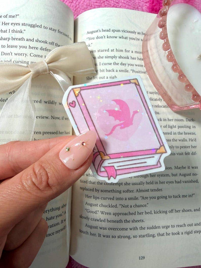 A hand with manicured nails holds a pink fairy-themed sticker or card in front of an open book, with a white ribbon and a pink beaded bracelet visible around it.