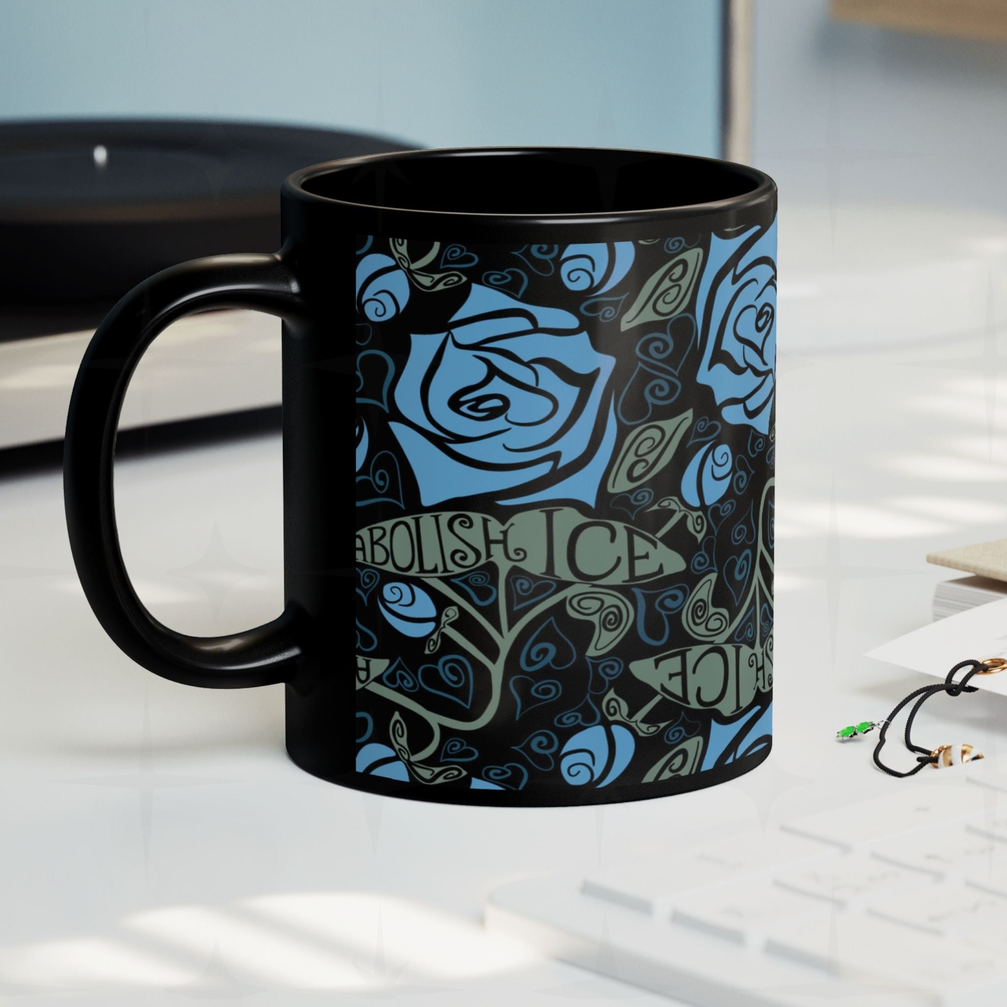 Abolish ICE black ceramic mug on a desk surface, gothic blue rose and hand-lettered activist wrap print, side view