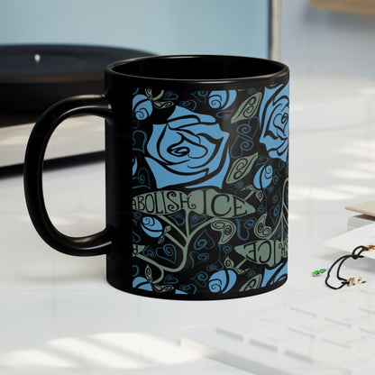 Abolish ICE black ceramic mug on a desk surface, gothic blue rose and hand-lettered activist wrap print, side view