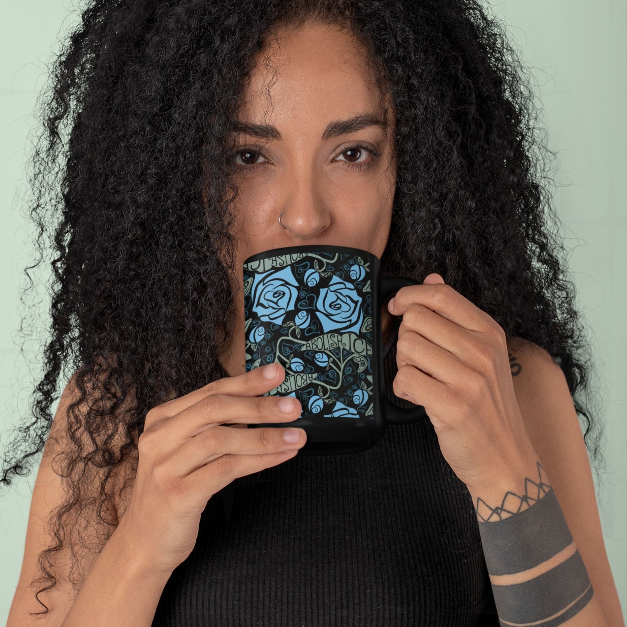 Woman with curly hair and nose ring drinking from the Abolish ICE black ceramic mug, gothic floral wrap print visible