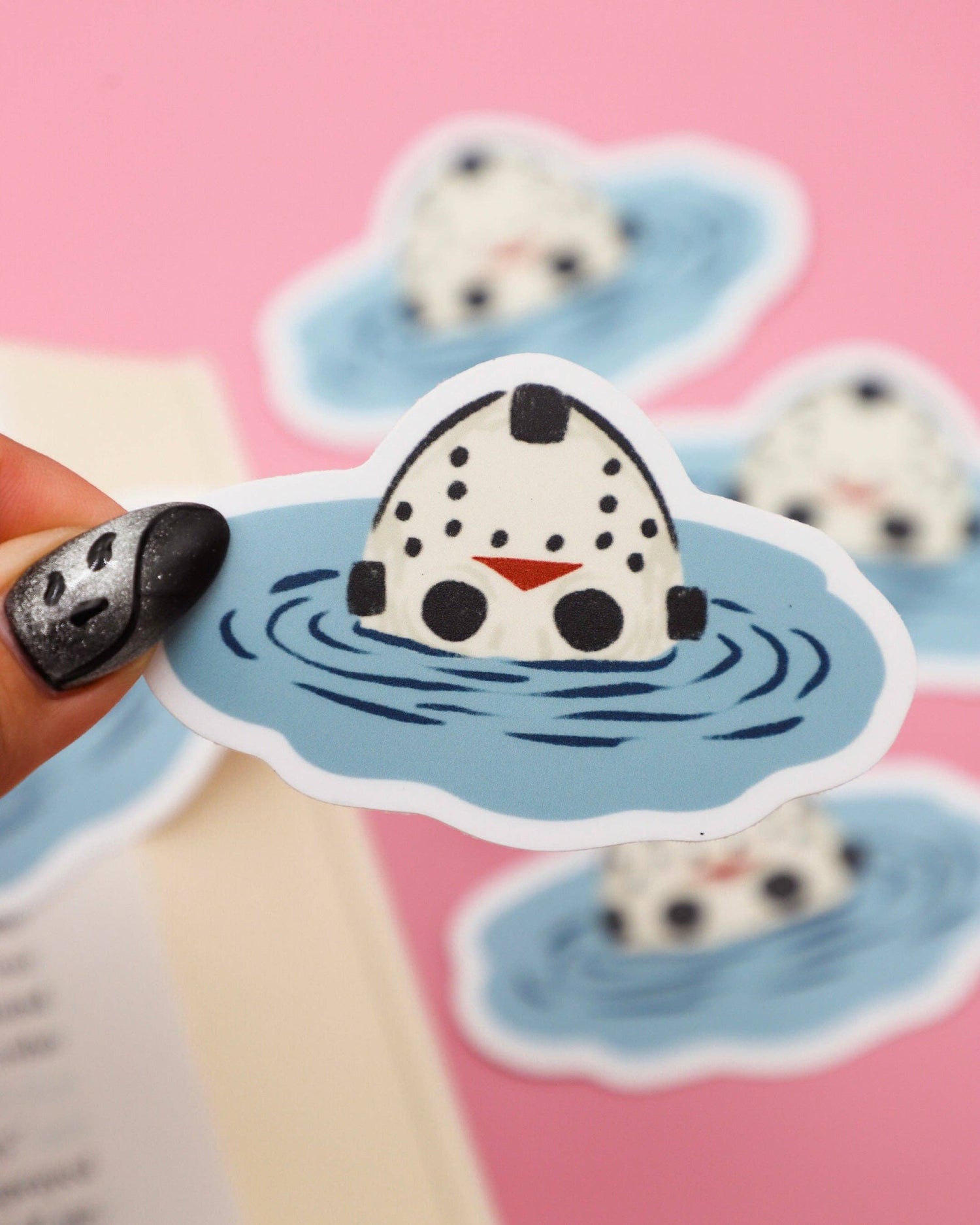 Sticker of a cartoon character in water held by a hand with black nail polish on a pink background.