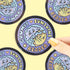 A circular sticker or coaster featuring a cartoon grumpy sunshine reader character, held by a hand with yellow nail polish against a pale yellow background with other copies of the sticker visible.