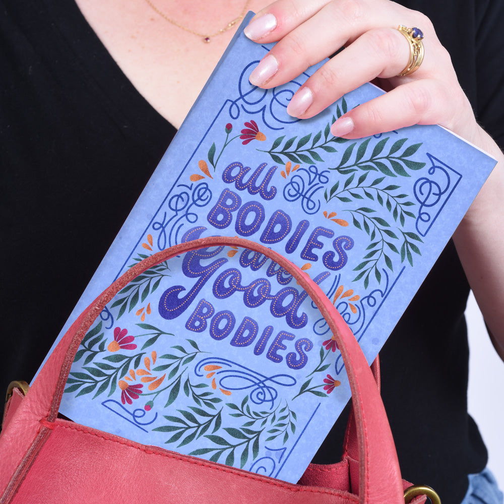 hands putting a blue notebook with flower illustrations and text "all bodies are good bodies" into a pink purse