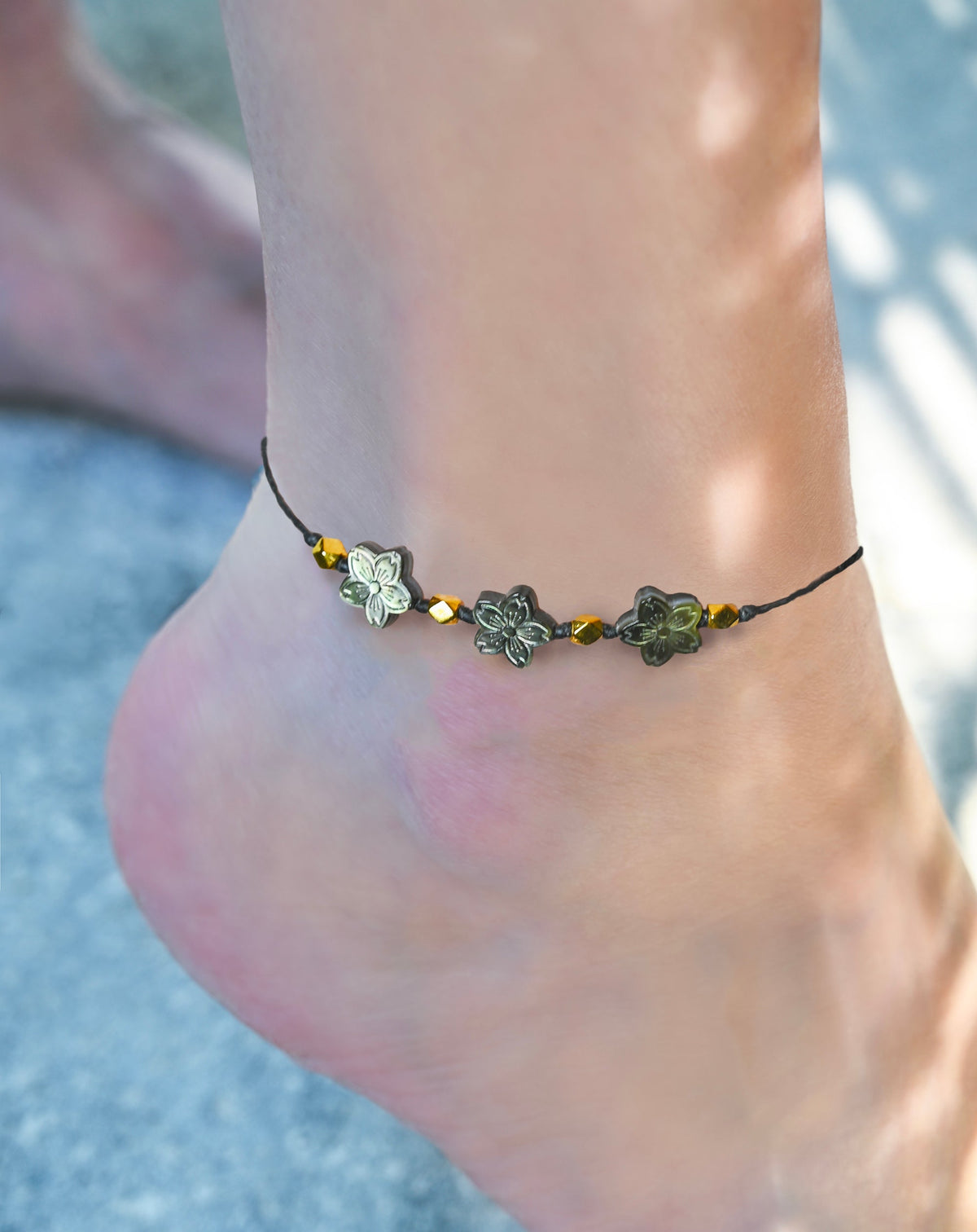 Close up of a black ankle bracelet with mother of pearl flowers and gold accents on a foot.