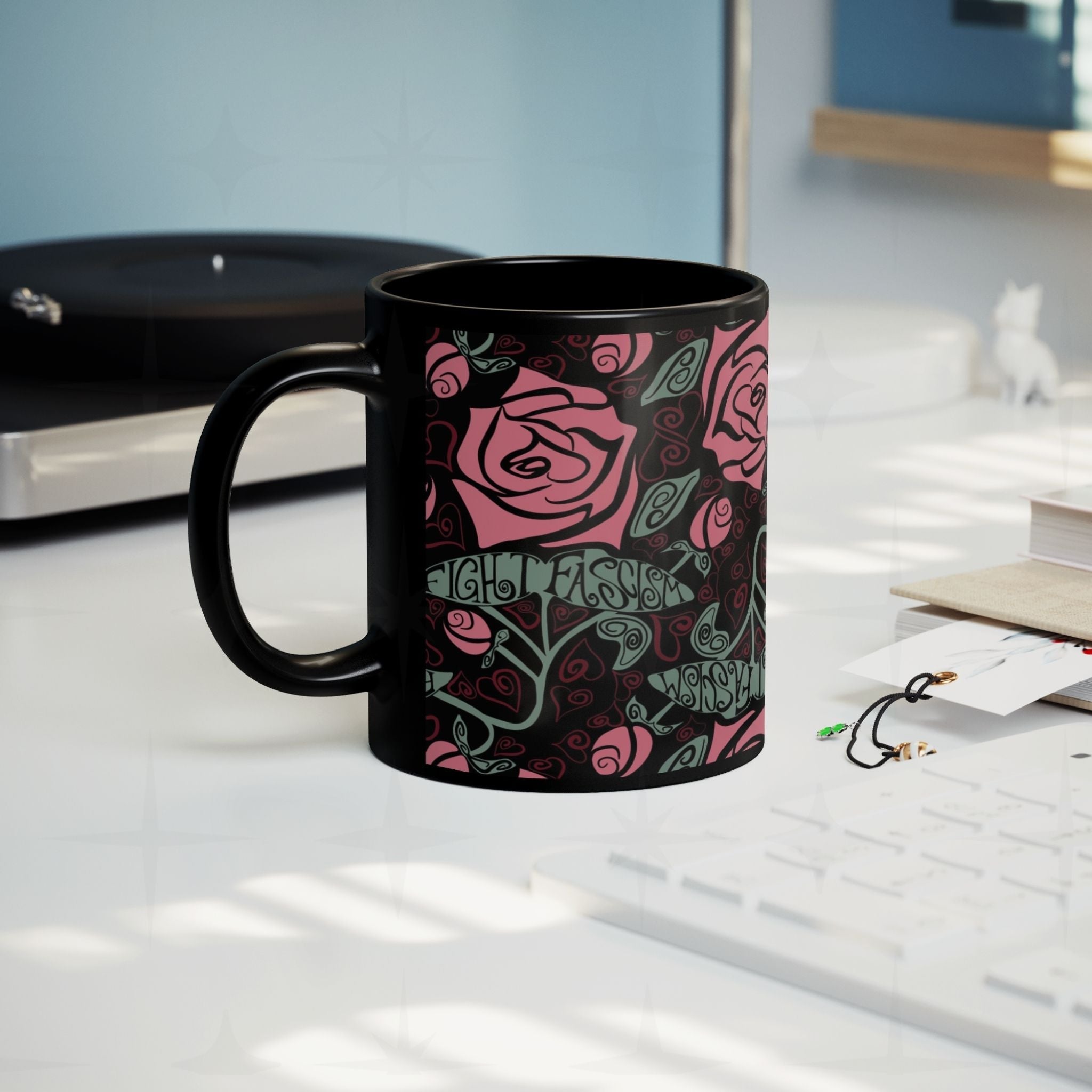 Fight Fascism anti-fascist coffee mug on a desk beside a record player, Art Nouveau rose pattern visible, cozy creative workspace lifestyle shot