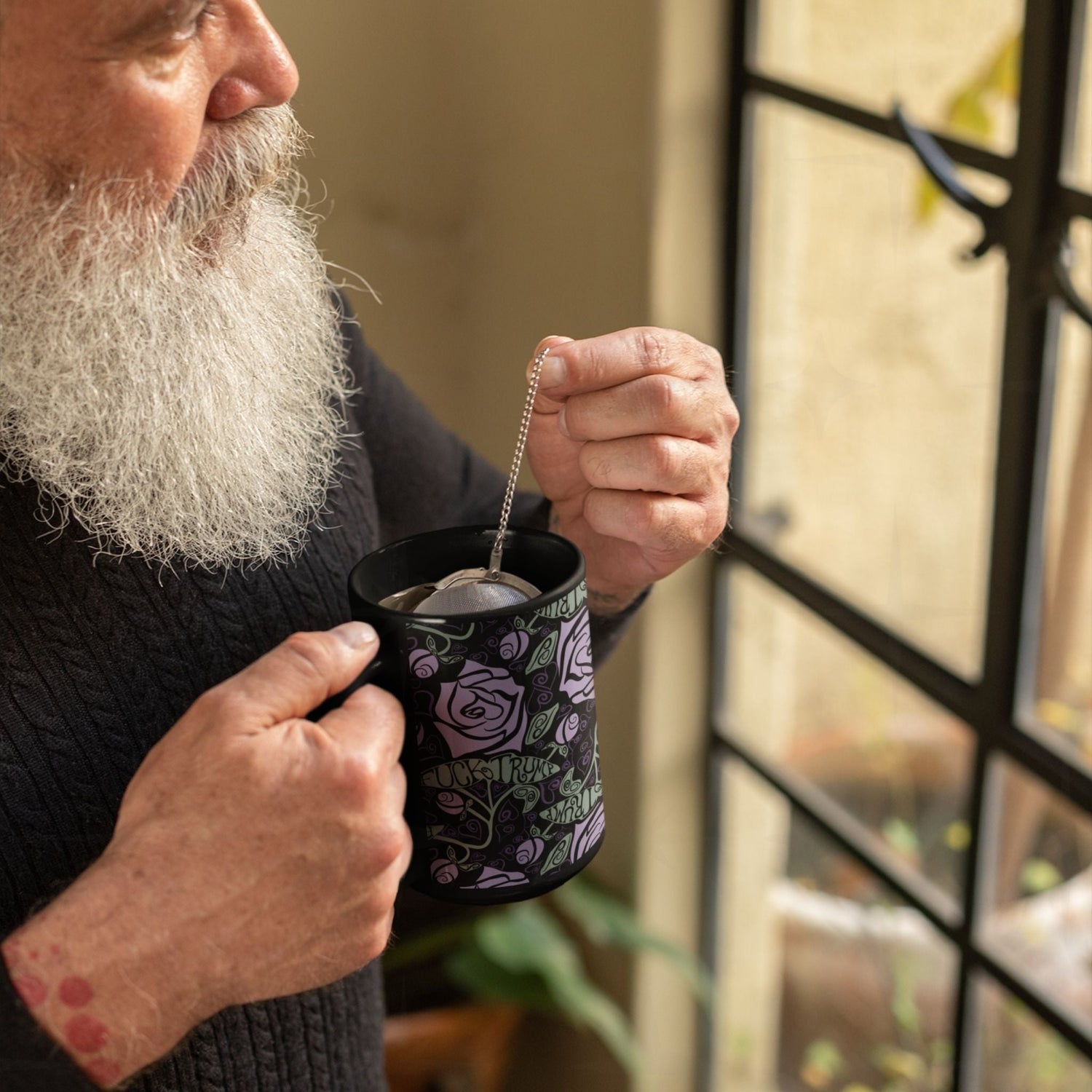 Older man with grey beard steeping tea in fuck Trump hidden message mug by a window