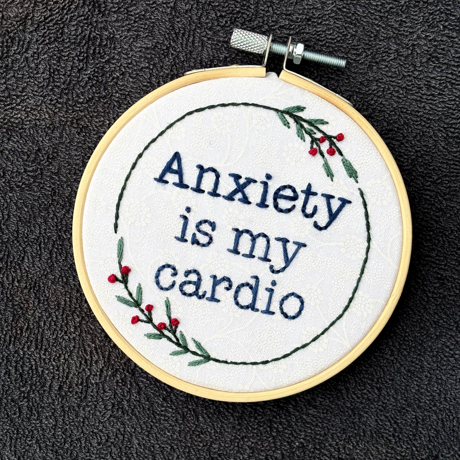 "Anxiety is my cardio." Quote hand embroidered in dark blue in typewriter font. Text is surrounded by minimalist floral wreath of dark green stems, green leaves, and red buds.. Fabric is white-on-white cotton. Displayed as wall art in a light wooden embroidery hoop.