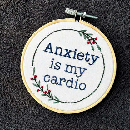 "Anxiety is my cardio." Quote hand embroidered in dark blue in typewriter font. Text is surrounded by minimalist floral wreath of dark green stems, green leaves, and red buds.. Fabric is white-on-white cotton. Displayed as wall art in a light wooden embroidery hoop.