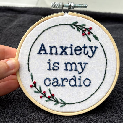 "Anxiety is my cardio." Quote hand embroidered in dark blue in typewriter font. Text is surrounded by minimalist floral wreath of dark green stems, green leaves, and red buds.. Fabric is white-on-white cotton. Displayed as wall art in a light wooden embroidery hoop.