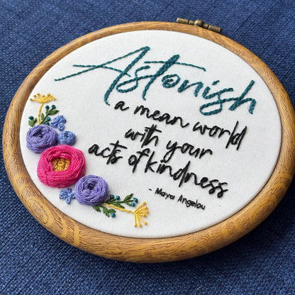 "Astonish a mean world with your acts of kindness. Maya Angelou." Astonish hand embroidered in dark teal. Remaining quote and attribution hand embroidered in black. In the lower left side, a cluster of flowers with bright pink and purple roses, golden pointy flowers, and green stems with leaves and dark teal flower buds. Art is displayed in a medium dark grained wood-look embroidery hoop.