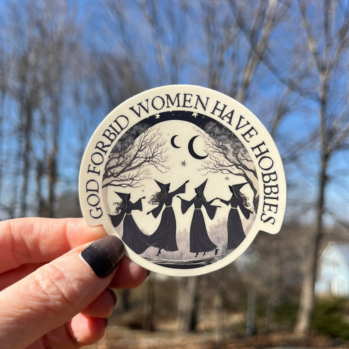 A hand with dark nail polish holds a round sticker outdoors against a blurred background of bare trees and blue sky. The sticker has a black and white illustration of three witches holding a staff under a crescent moon and stars, with the text &