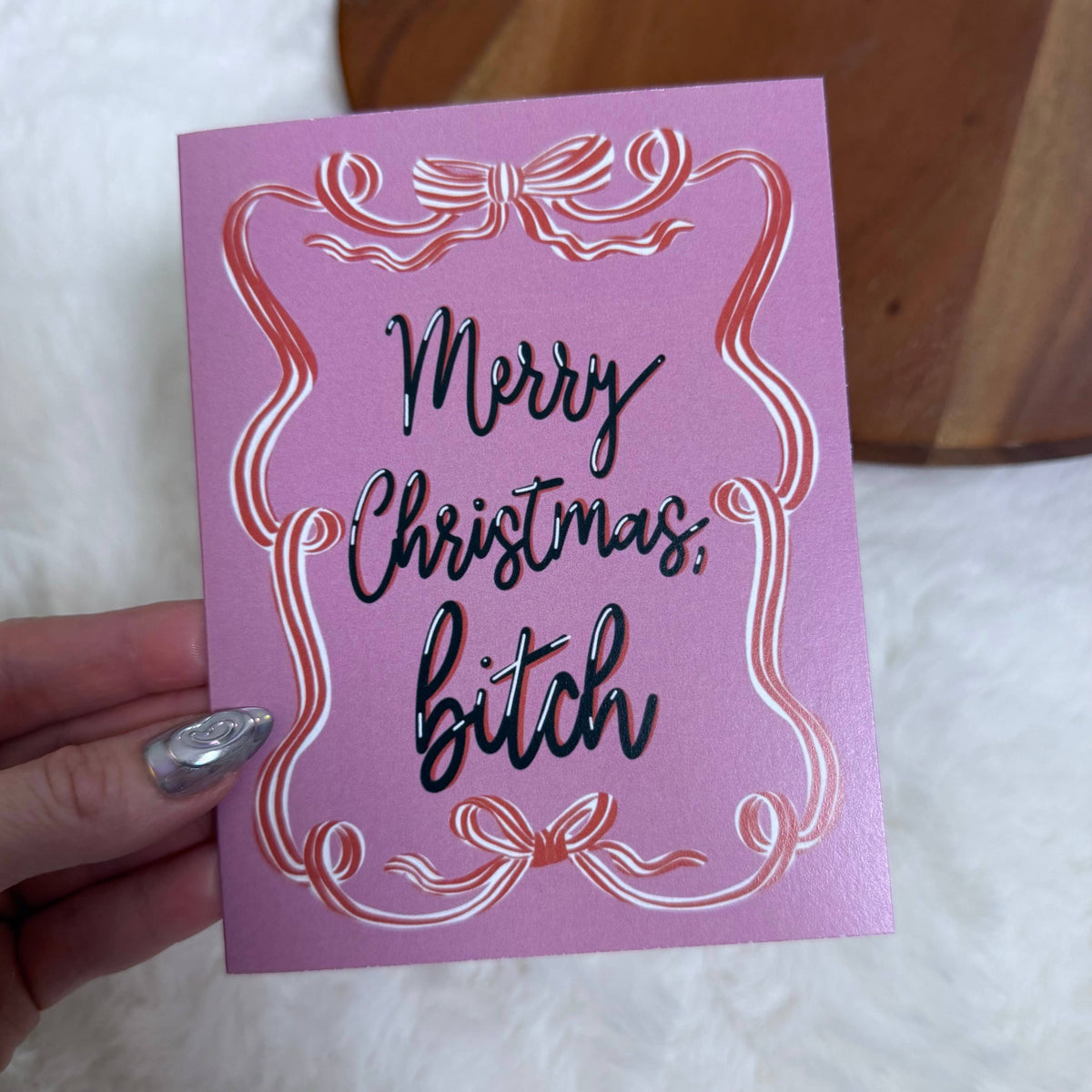 A hand holding a pink Christmas greeting card with metallic rose-gold foil scrollwork and black cursive text reading &
