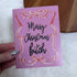 A hand holding a pink Christmas greeting card with metallic rose-gold foil scrollwork and black cursive text reading &