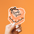 A hand with white manicured nails holds a scalloped die-cut sticker that reads &