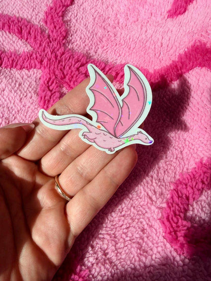 A hand holds a pink dragon-shaped sticker or patch with iridescent glitter accents, set against a textured hot pink blanket.