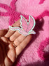 A hand holds a pink dragon-shaped sticker or patch with iridescent glitter accents, set against a textured hot pink blanket.
