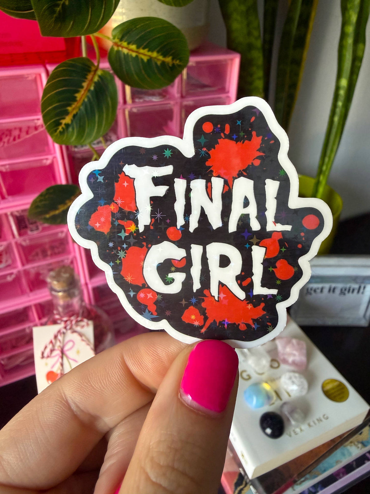 A hand with bright pink nail polish holds a die-cut sticker that reads &