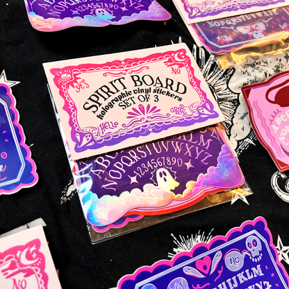 A set of three holographic vinyl stickers featuring gothic designs, including a Spirit Board, an Alphabet Board, and a Numbers Board, displayed on a black background with other stickers.