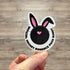 Sticker with bunny ears and text held by a hand on a wooden surface
