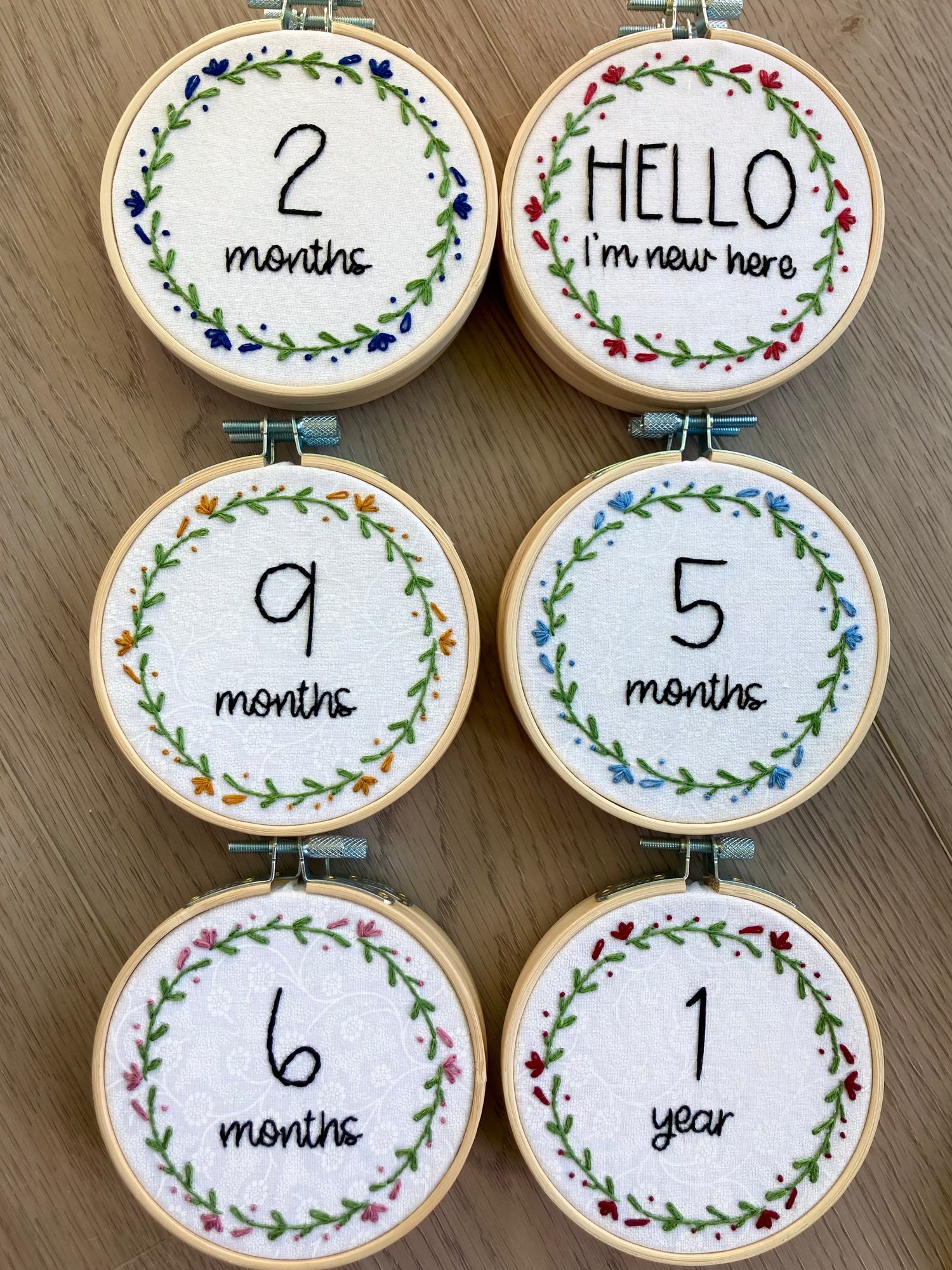 Collection of completed 4-inch wood embroidery hoops with baby milestone text surrounded by simple flower wreaths. Hello, I&