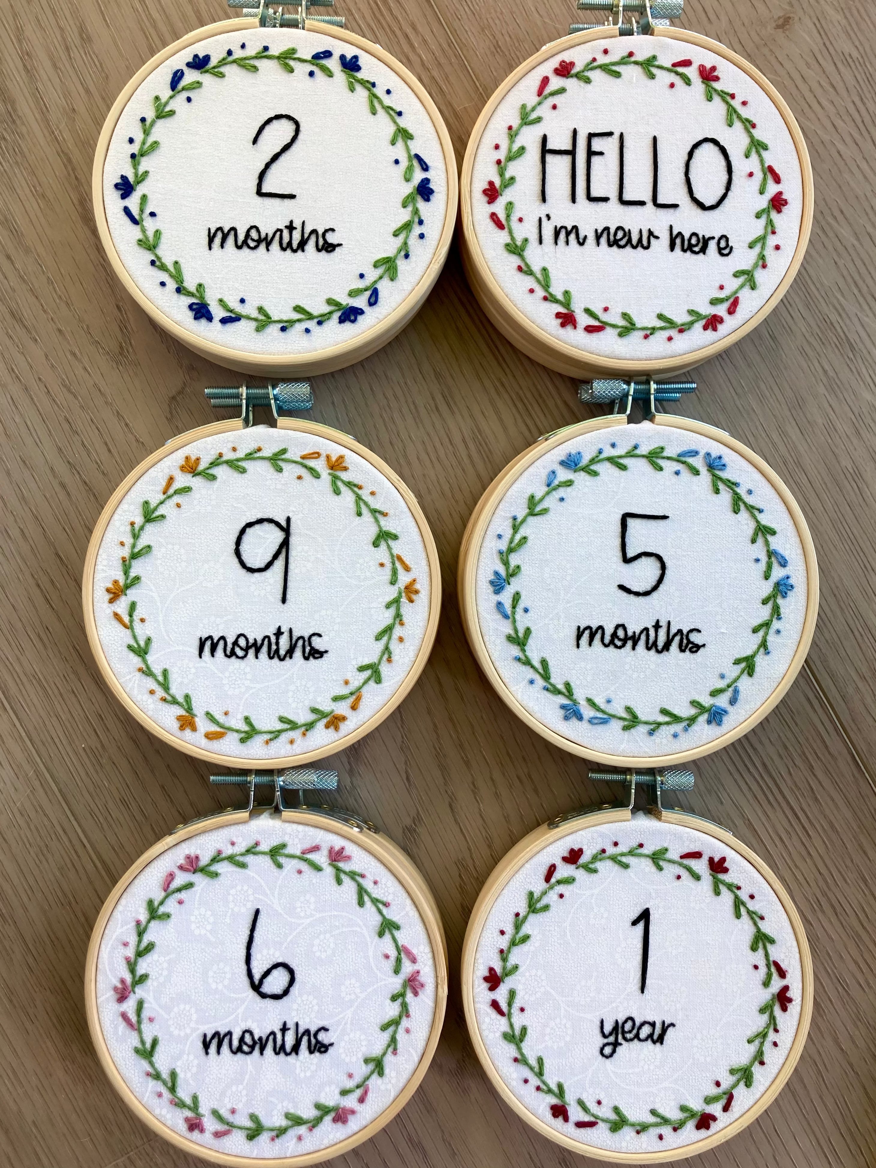 Collection of completed 4-inch wood embroidery hoops with baby milestone text surrounded by simple flower wreaths. Hello, I&