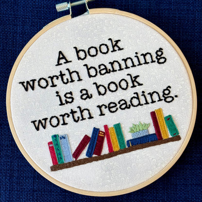"A book worth banning is a book worth reading". Quote hand embroidered in black serif font. Below the text is an embroidered bookshelf with several different colored books, as well as a light blue planter holding a light green plant. Sewn on white-on-white cotton. Framed in a light wood embroidery hoop as wall art or bookshelf art.