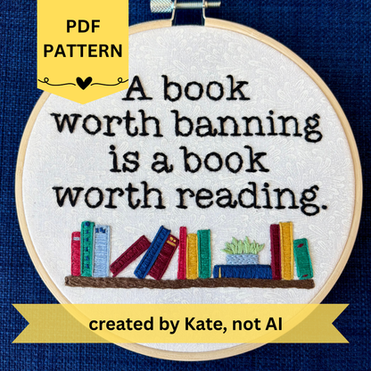 PDF pattern created by Kate, not AI. "A book worth banning is a book worth reading". Quote hand embroidered in black serif font. Below the text is an embroidered bookshelf with several different colored books, as well as a light blue planter holding a light green plant. Sewn on white-on-white cotton. Framed in a light wood embroidery hoop as wall art or bookshelf art.