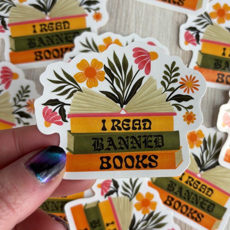 hand holding a sticker of a stack of books that says "I read banned books" with flowers