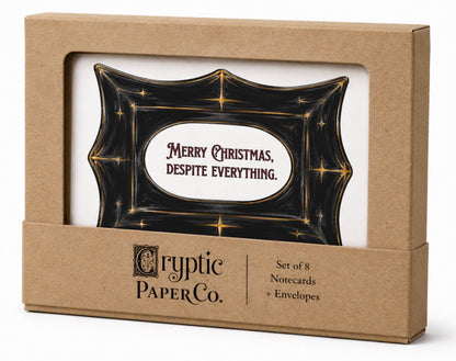 Cryptic Paper Co. Christmas greeting card set in a kraft paper box with a clear window, featuring a black card with gold stars and the text &