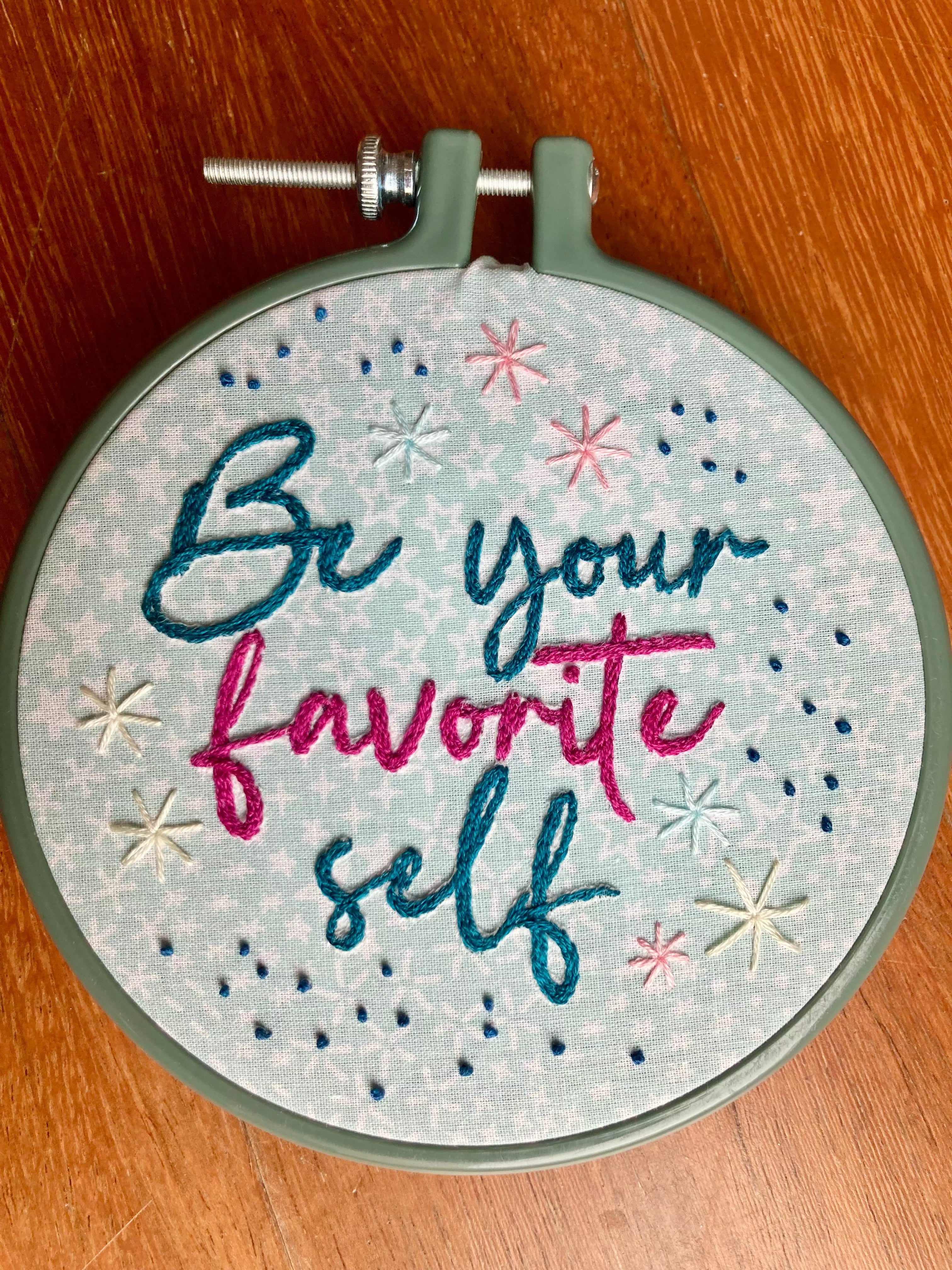 Be Your Favorite Self is hand embroidered in a script font on light blue cotton printed with white stars. Favorite is sewn in bright pink, be your and self are stitched in dark teal, and stars surround the quote. This wall art is framed in a blue plastic embroidery frame.