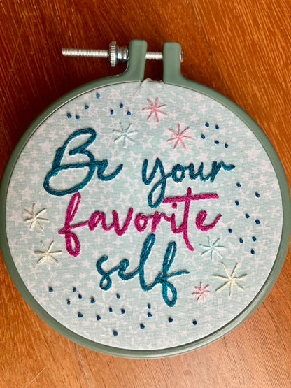 Be Your Favorite Self is hand embroidered in a script font on light blue cotton printed with white stars. Favorite is sewn in bright pink, be your and self are stitched in dark teal, and stars surround the quote. This wall art is framed in a blue plastic embroidery frame.
