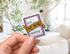 A hand holds a die-cut sticker shaped like a book chapter, featuring a purple border, white page background, and a gold ribbon banner that reads &