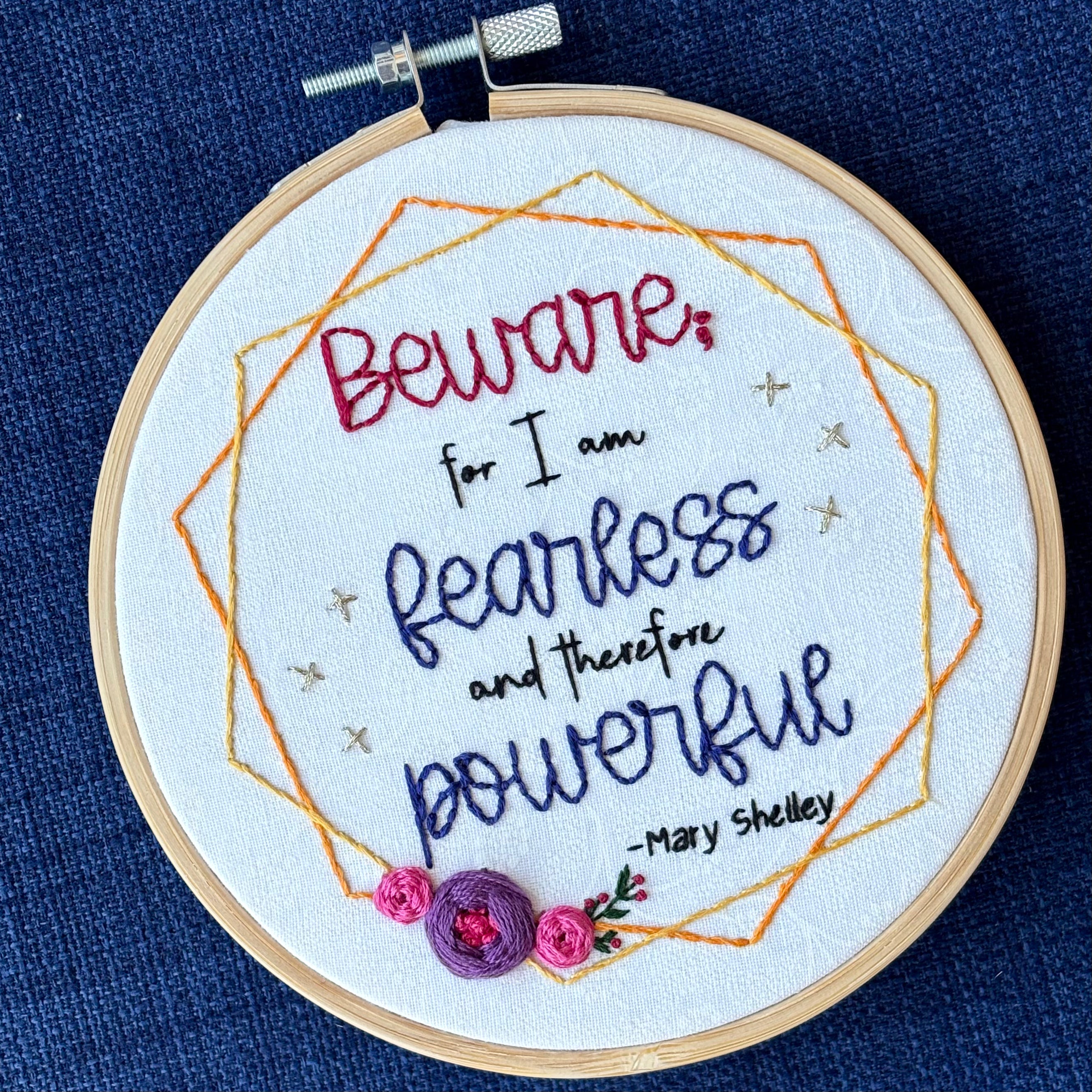 "Beware; for I am fearless and therefore powerful - Mary Shelley". Quote hand embroidered in dark red, black, and deep royal blue. Framed in yellow and orange hexagon wreaths with floral bundle on the bottom left. Gold stars sparkle on the sides. Fabric is white-on-white cotton. Displayed as wall art in a light wooden embroidery hoop.