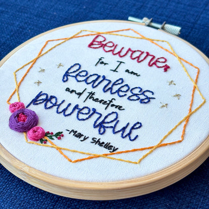 "Beware; for I am fearless and therefore powerful - Mary Shelley". Quote hand embroidered in dark red, black, and deep royal blue. Framed in yellow and orange hexagon wreaths with floral bundle on the bottom left. Gold stars sparkle on the sides. Fabric is white-on-white cotton. Displayed as wall art in a light wooden embroidery hoop.