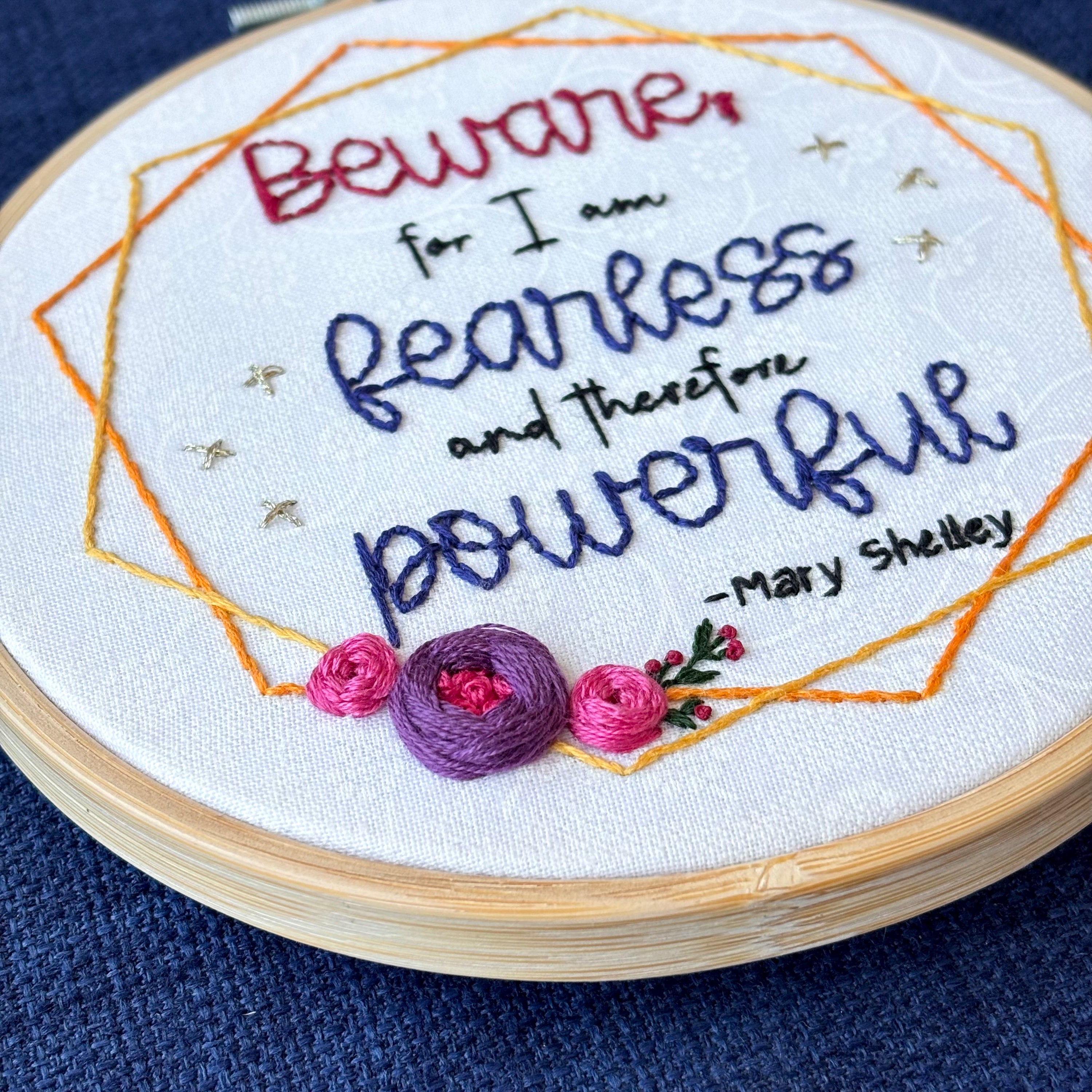 "Beware; for I am fearless and therefore powerful - Mary Shelley". Quote hand embroidered in dark red, black, and deep royal blue. Framed in yellow and orange hexagon wreaths with floral bundle on the bottom left. Gold stars sparkle on the sides. Fabric is white-on-white cotton. Displayed as wall art in a light wooden embroidery hoop.