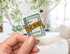 A hand holds a die-cut sticker shaped like a book cover with the text &