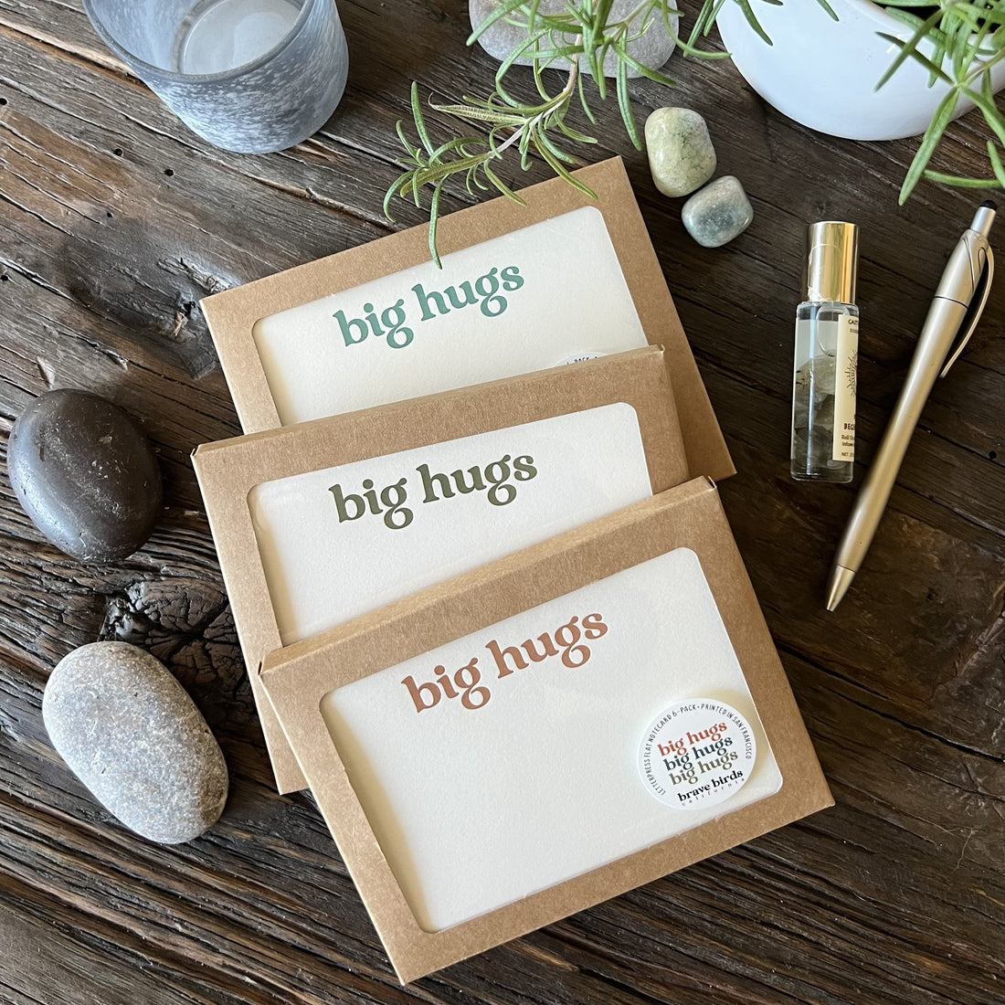 Three craft paper boxes with clear windows showing printed notecards with the words "big hugs" in teal, olive and warm orange ink. Styled on a wooden table with river rocks, a gold pen, a bottle of essential oils and a small peak of rosemary branches. 