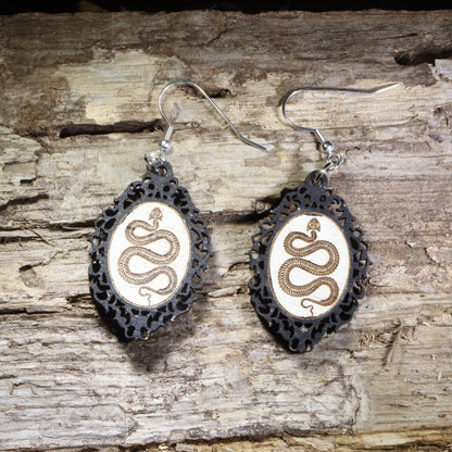 Handcrafted black framed snake wooden dangle earrings featuring a detailed serpent coiled within an ornate gothic-inspired frame. Lightweight laser-cut wooden earrings with hypoallergenic .925 sterling silver hooks, perfect for snake lovers, gothic fashion, reptile enthusiasts, witchy style, dark academia, and nature-inspired handmade jewelry.