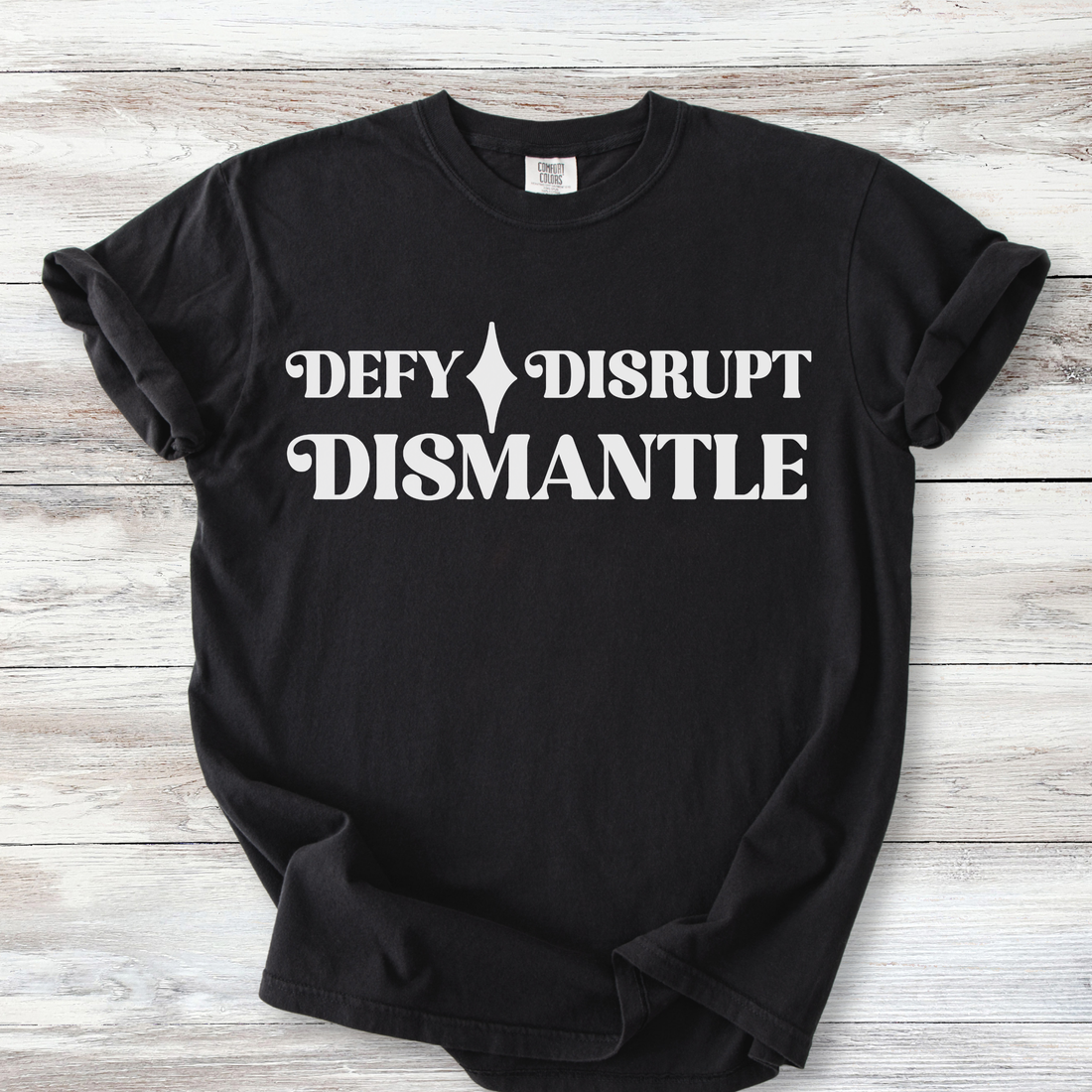 The words DEFY DISRUPT DISMANTLE.  DEFY AND DISRUPT are on the top line with a star between them.  DISMANTLE is the bottom line, lining up vertically with the top line. Lettering is a stylized retro style font.  