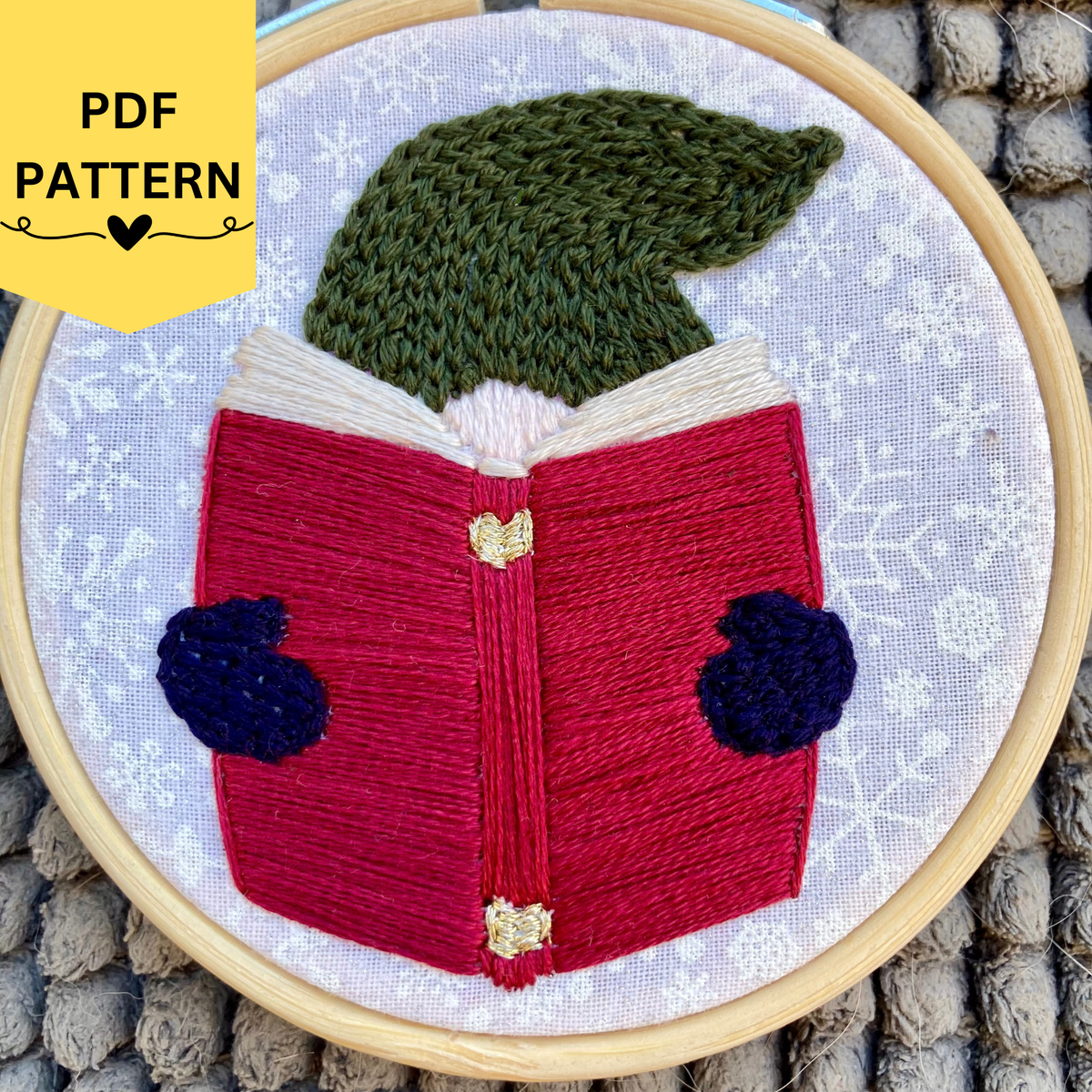 PDF pattern. Hand embroidered Christmas gnome with a pine green knit winter hat reading a red book, wearing navy blue mittens. Stitched on white cotton with sparkly white snowflakes printed on the fabric. Displayed in light wooden hoop as a DIY Christmas ornament.