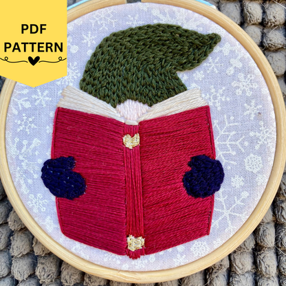 PDF pattern. Hand embroidered Christmas gnome with a pine green knit winter hat reading a red book, wearing navy blue mittens. Stitched on white cotton with sparkly white snowflakes printed on the fabric. Displayed in light wooden hoop as a DIY Christmas ornament.