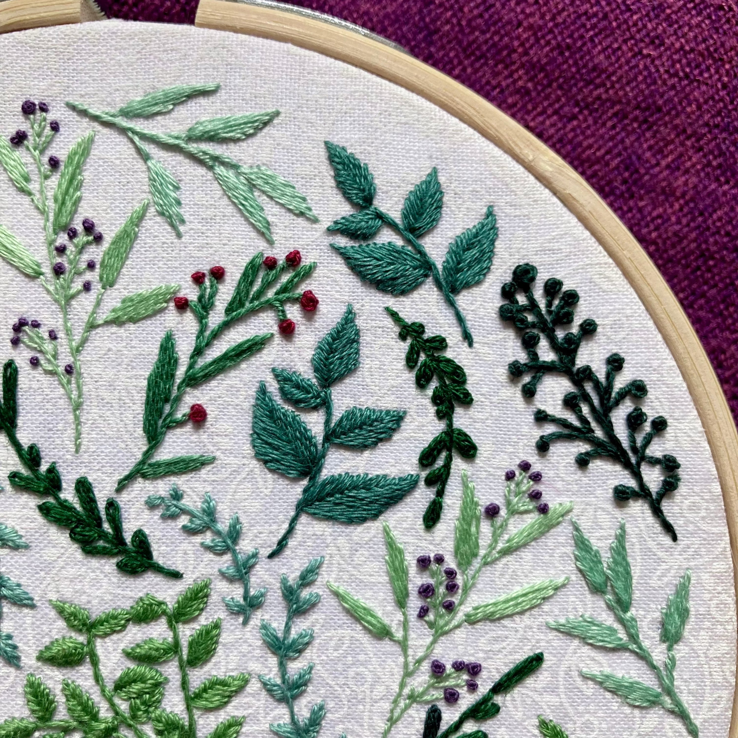 8 different botanicals hand embroidered in a scattered pattern on white cotton fabric. There is a range of yellow green and blue green leaves and stems, as well as red and purple flower buds. This wall art is framed in a light wood embroidery frame.