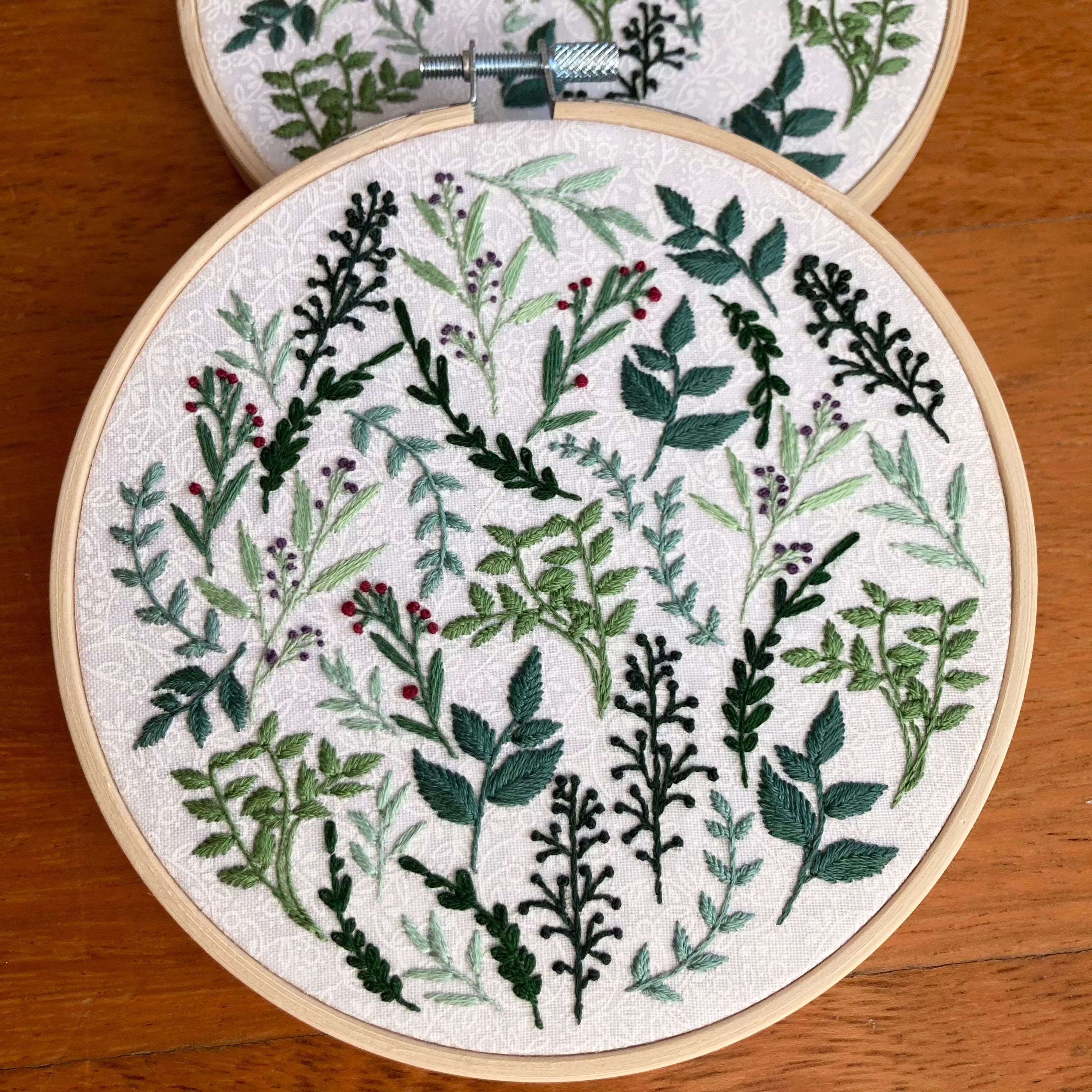 8 different botanicals hand embroidered in a scattered pattern on white cotton fabric. There is a range of yellow green and blue green leaves and stems, as well as red and purple flower buds. This wall art is framed in a light wood embroidery frame.
