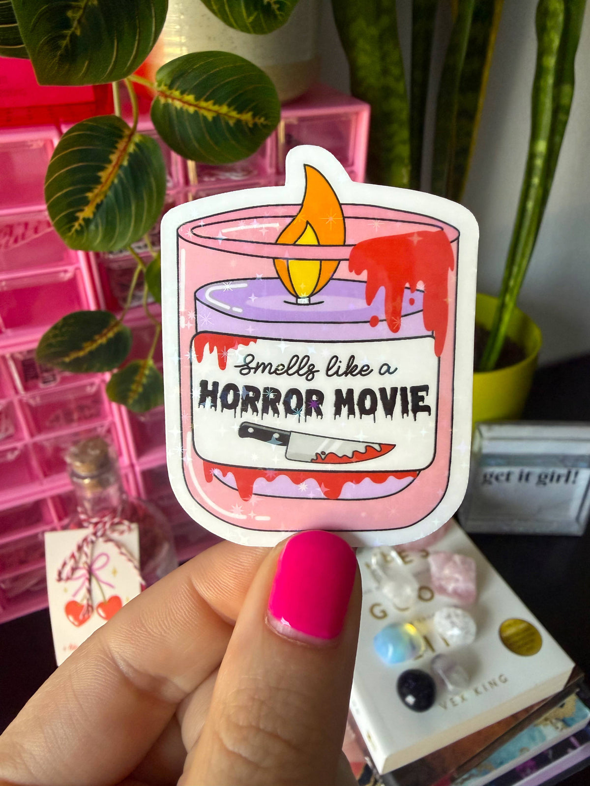 A hand with bright pink nail polish holds a die-cut sticker featuring a pink candle with dripping red wax, a yellow flame, and the text &